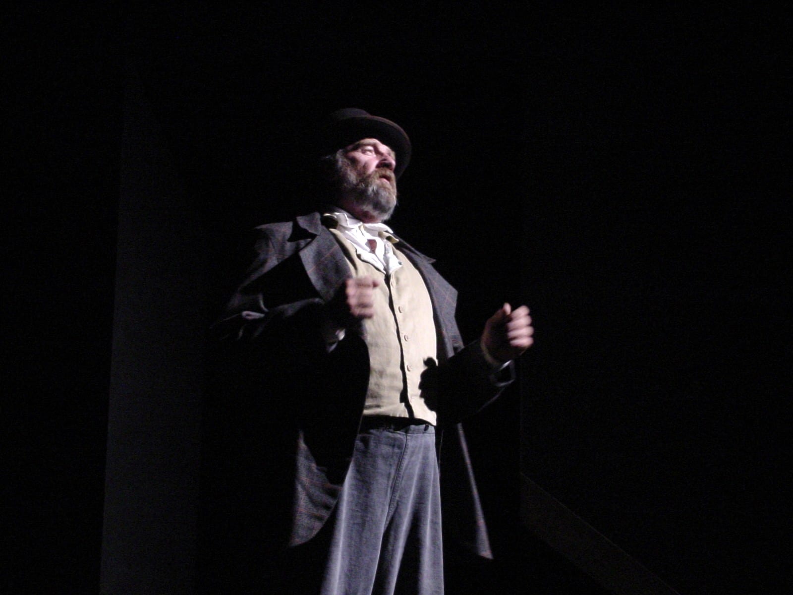 Bearded character in coat and bowler hat under stage light